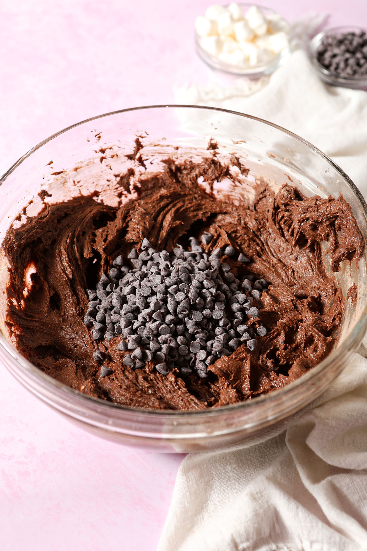 Chocolate chips sit on top of chocolate cookie dough on a pink surface next to a white linen