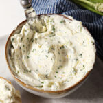 A bowl of jalapeño cream cheese on a grey plate with a knife in it on marble surface next to a blue linen