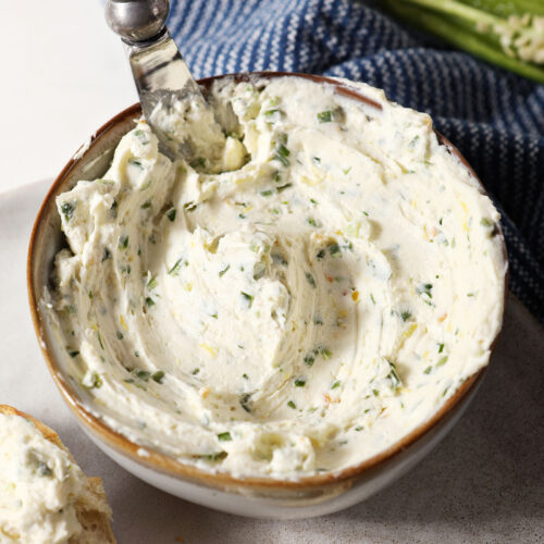 A bowl of jalapeño cream cheese on a grey plate with a knife in it on marble surface next to a blue linen