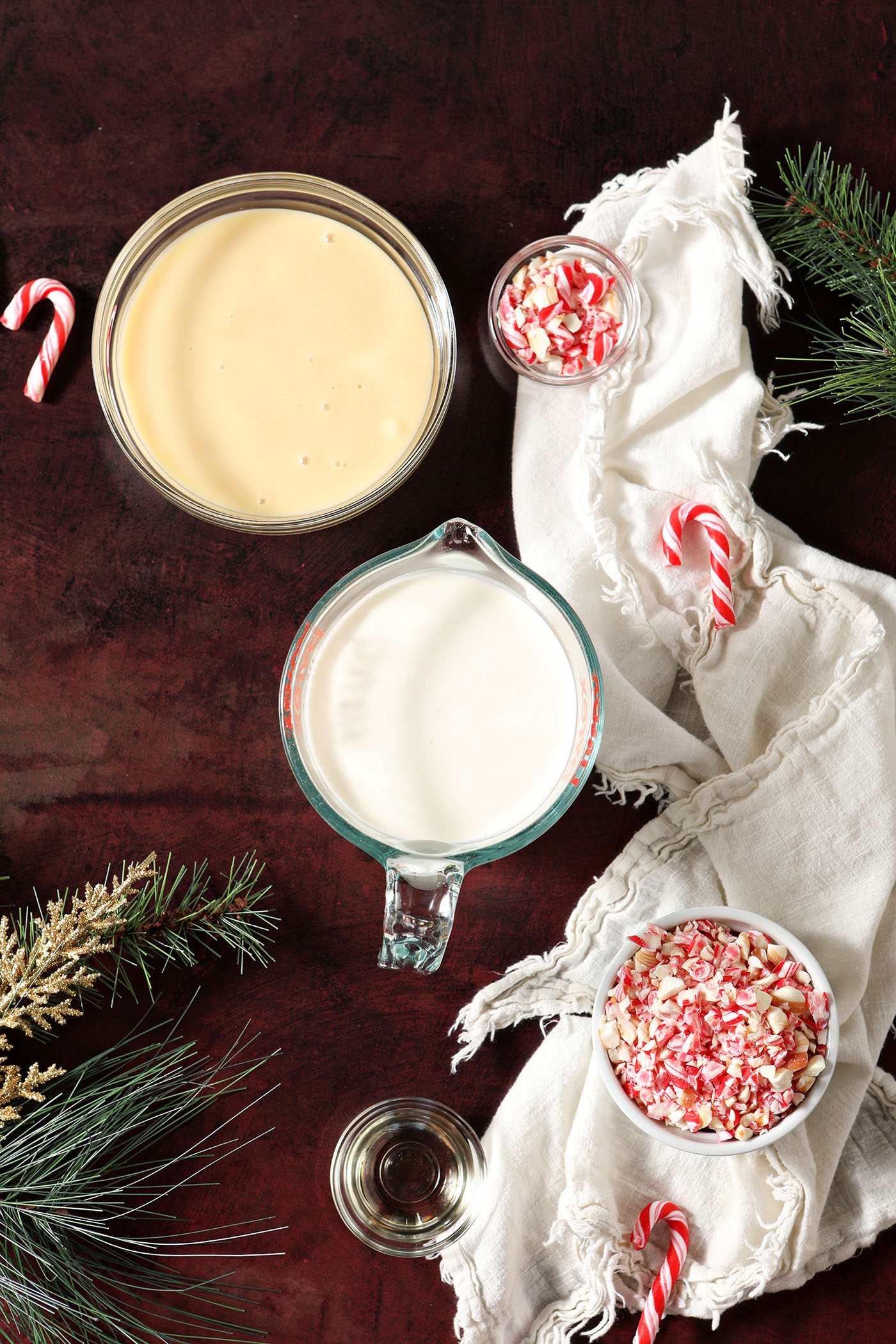 Ingredients to make no churn peppermint stick ice cream in bowls on a dark red surface next to a white linen and evergreen