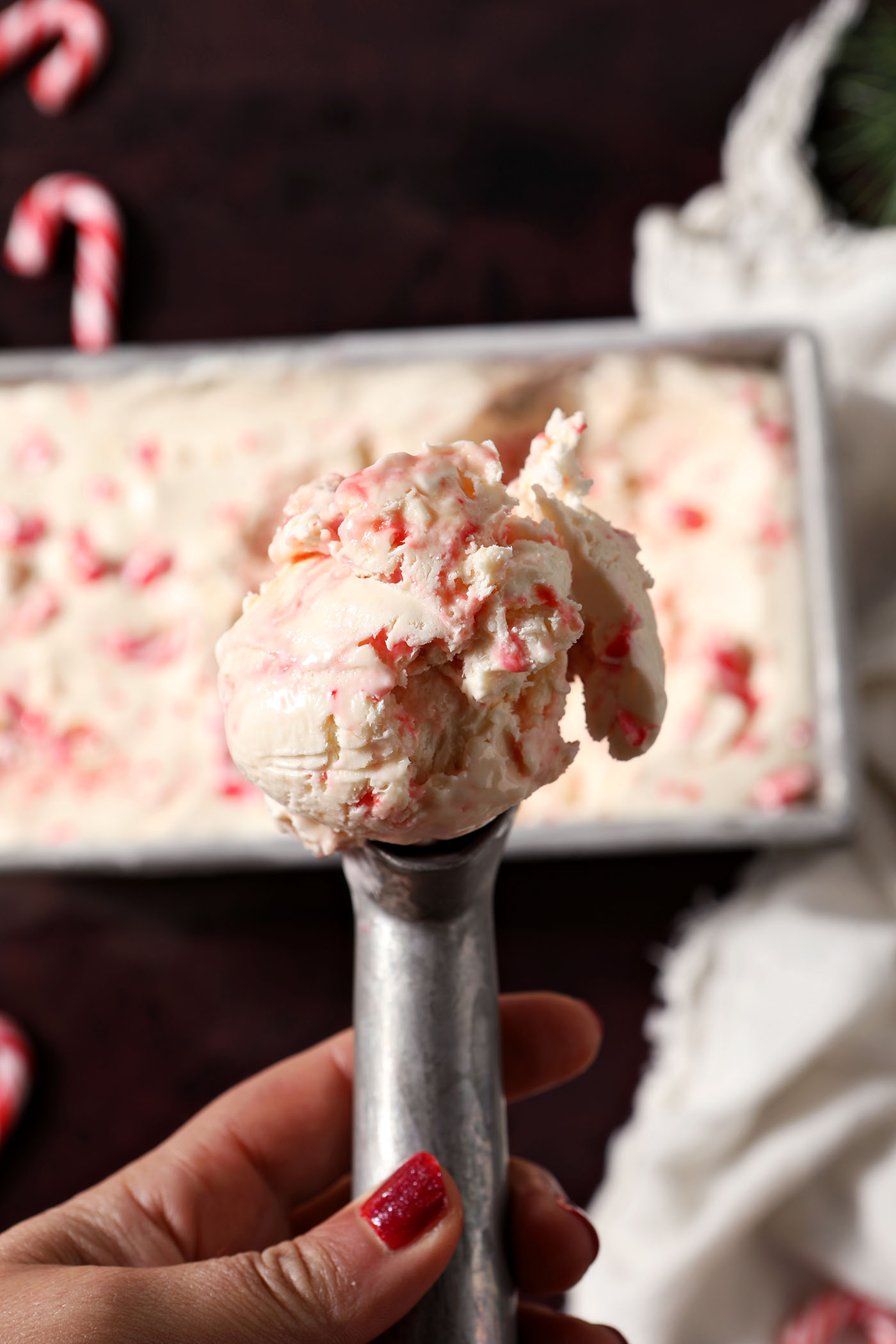 An ice cream scooper holds a scoop of no churn peppermint stick ice cream above a loaf pan on a dark red surface