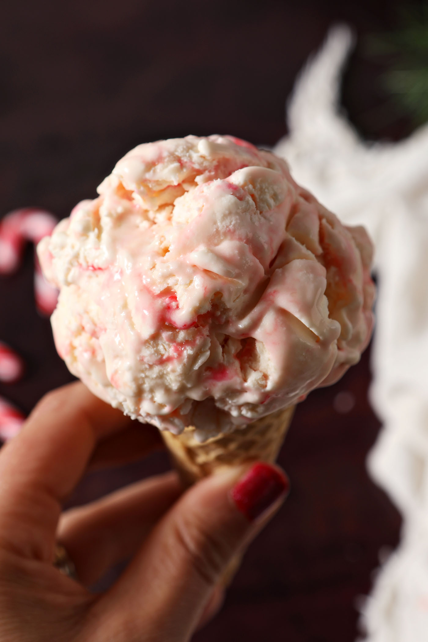 Overhead of a hand holding a cone with a scoop of No Churn Peppermint Ice Cream in it above a dark red surface