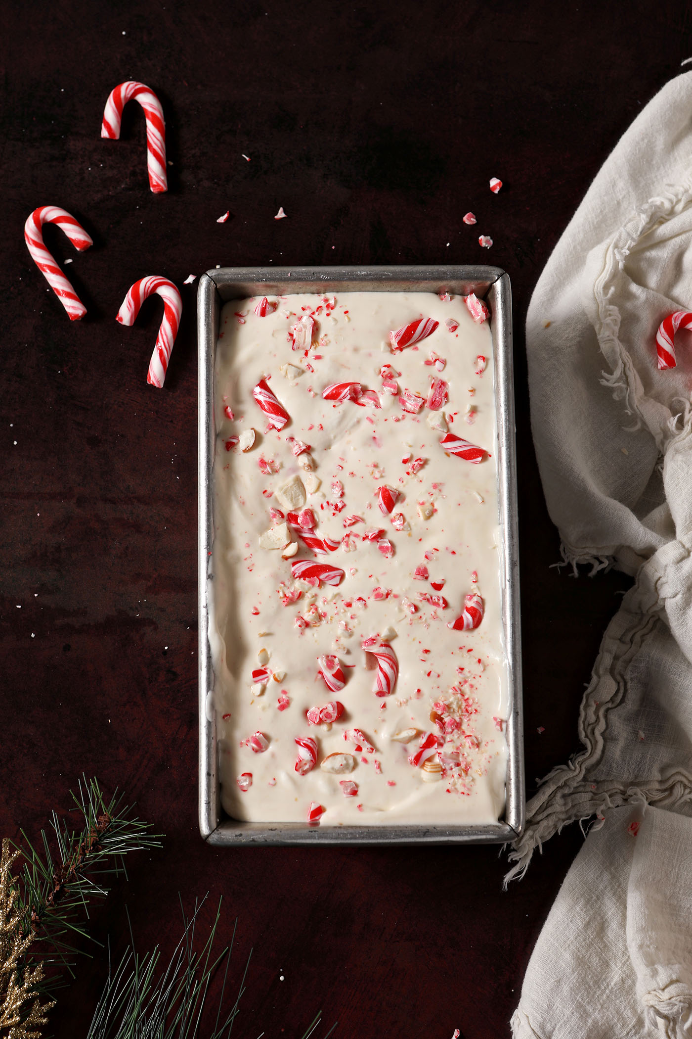No Churn Peppermint Ice Cream garnished with crushed candy canes and peppermints in a loaf pan on a dark red surface, after freezing