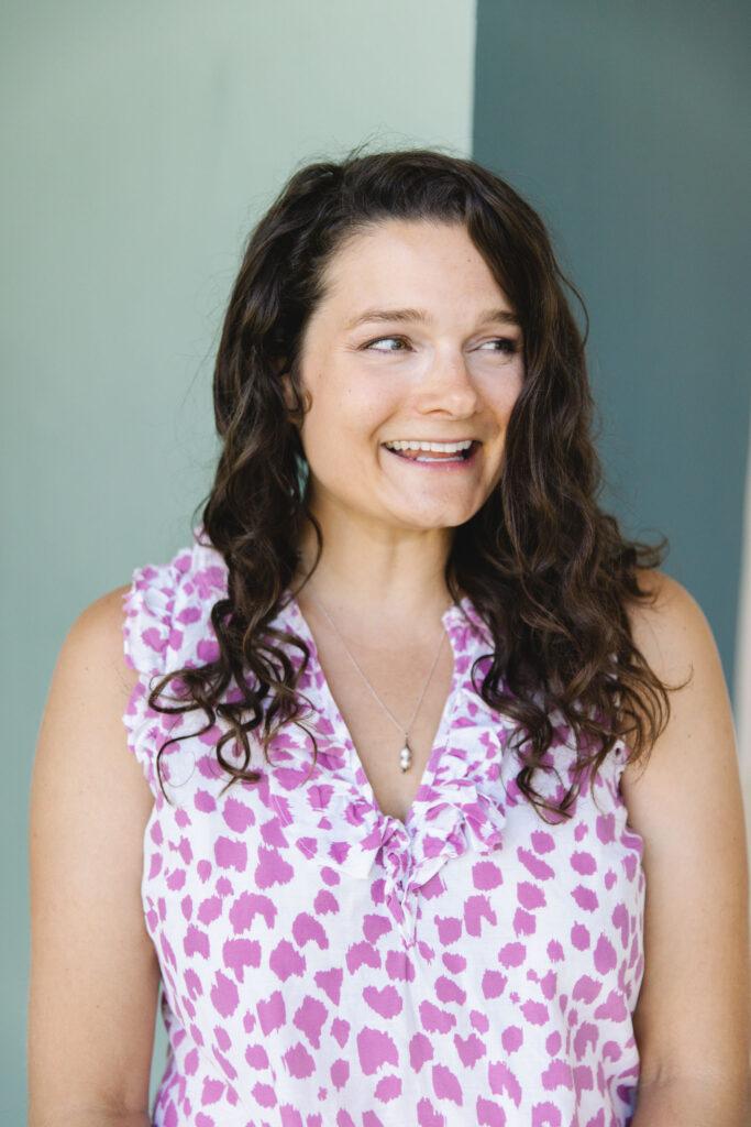 a light-skinned woman with dark brown hair wearing a purple and white polka dot shirt looks off to her left while smiling