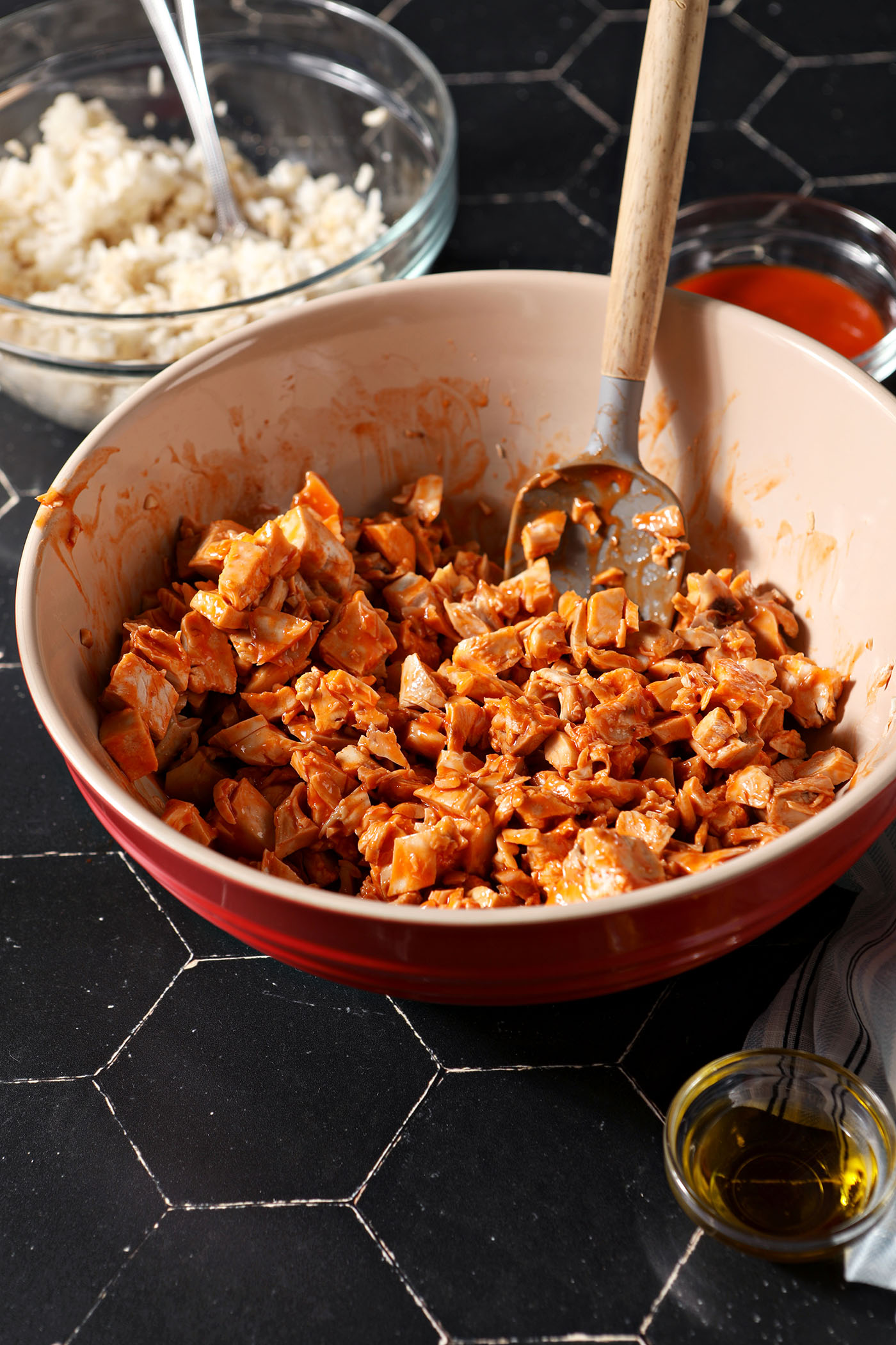 A red bowl of chopped chicken tossed in buffalo sauce on a black tiled surface