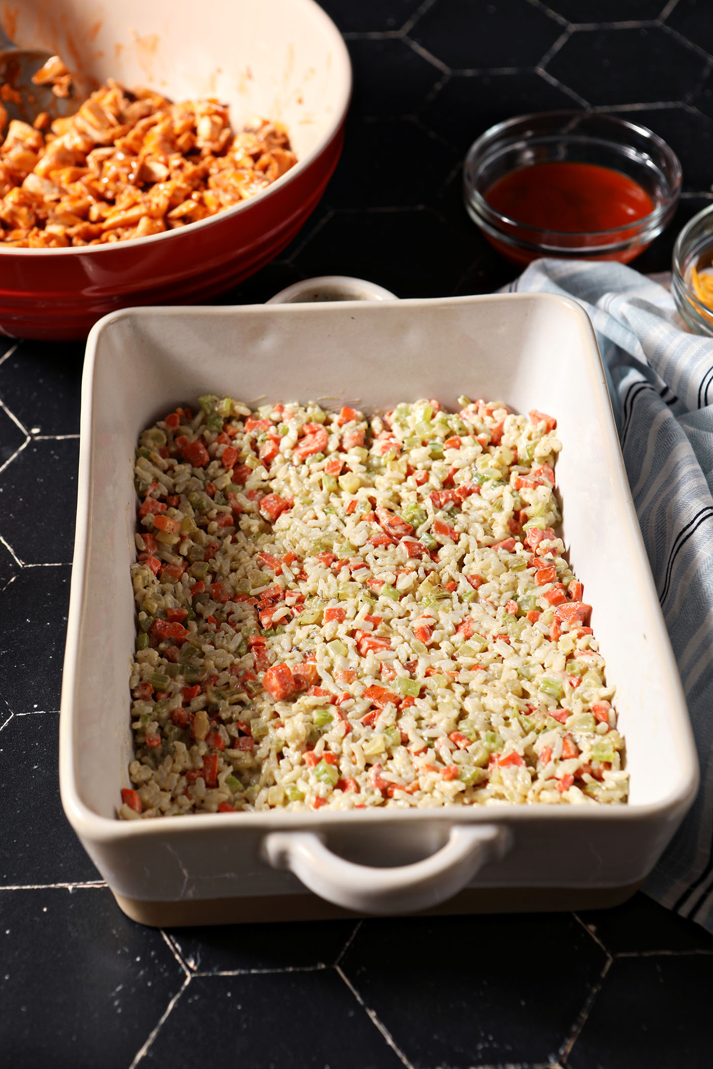 A layer of rice and veggies in the bottom of a casserole dish on a black tiled surface next to a bowl of saucy chicken
