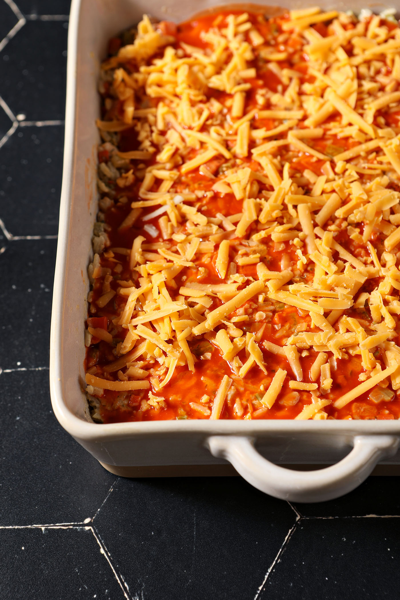 A casserole dish of Buffalo Chicken Casserole topped with shredded cheese on a black tiled surface before baking