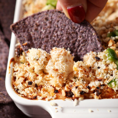 Close up of a hand dipping a blue corn tortilla chip into baked jalapeño popper dip