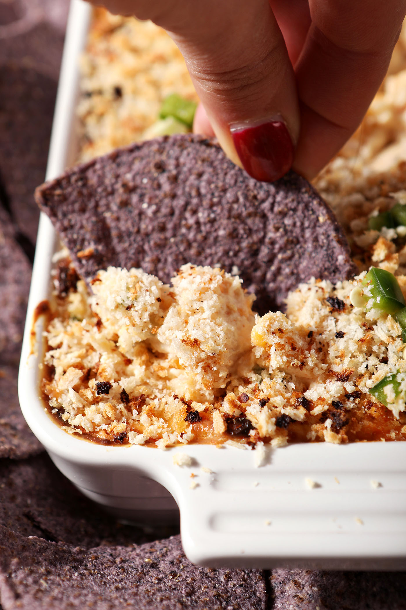 Close up of a hand dipping a blue corn tortilla chip into baked jalape&ntilde;o popper dip