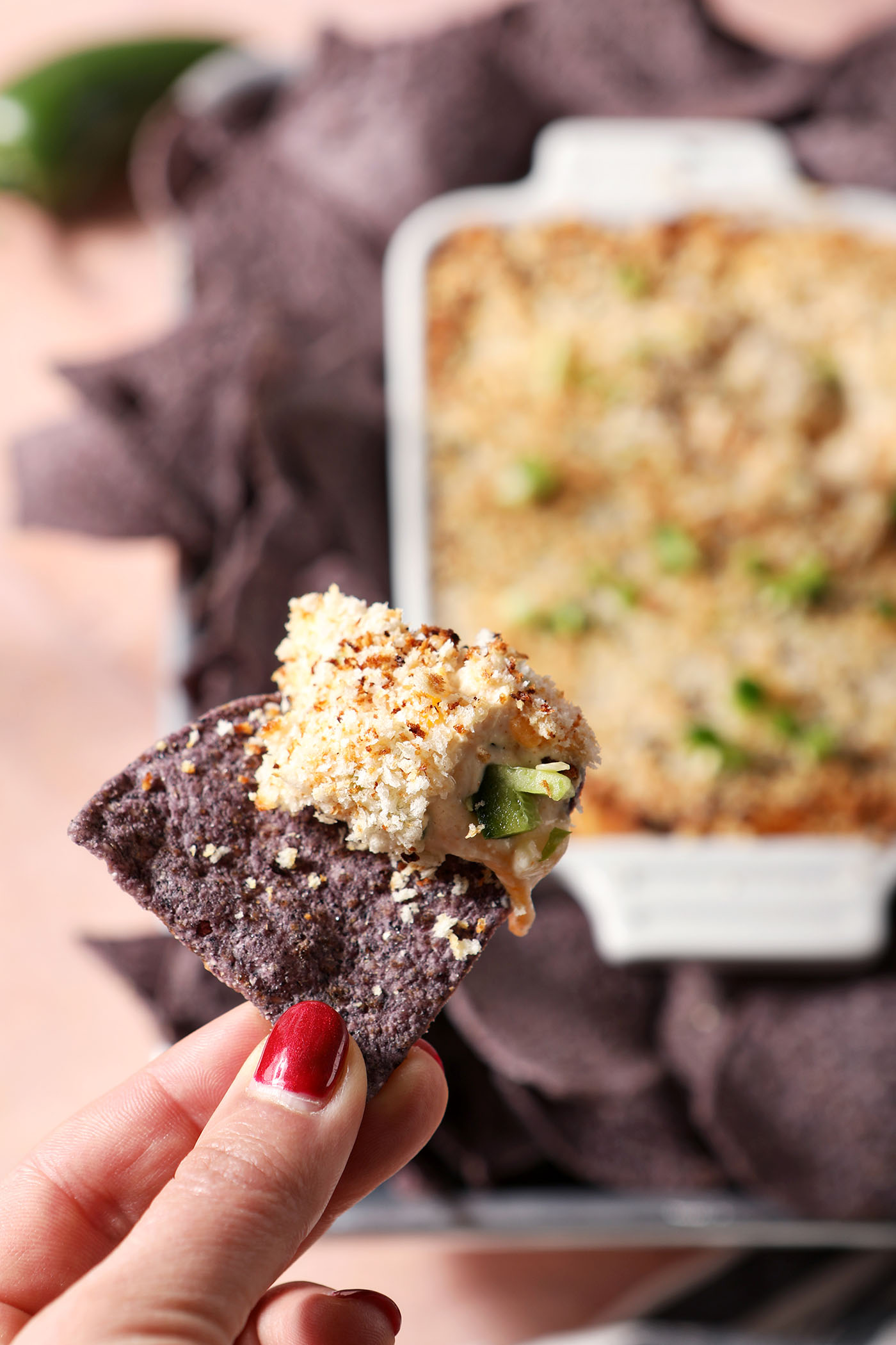 A hand holds a blue corn tortilla chip above a platter holding a cheesy baked dip and more tortilla chips