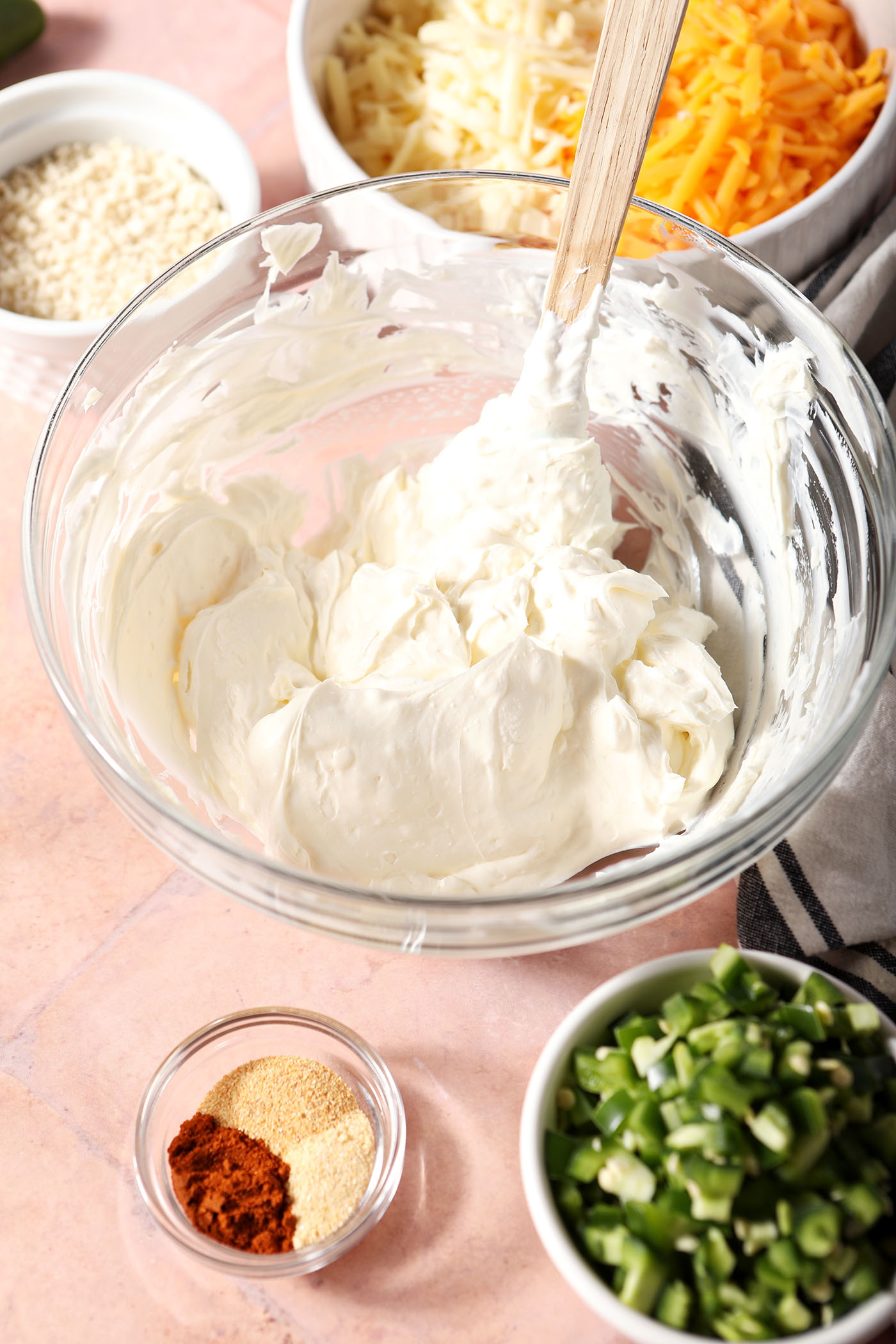 Cream cheese and sour cream, mixed in a bowl with a small rubber spatula, on a terracotta surface and surrounded by bowls of other ingredients