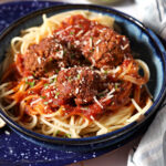 Side angle of a blue bowl holding a large serving of mom’s spaghetti and meatballs, garnished with parsley and parmesan cheese