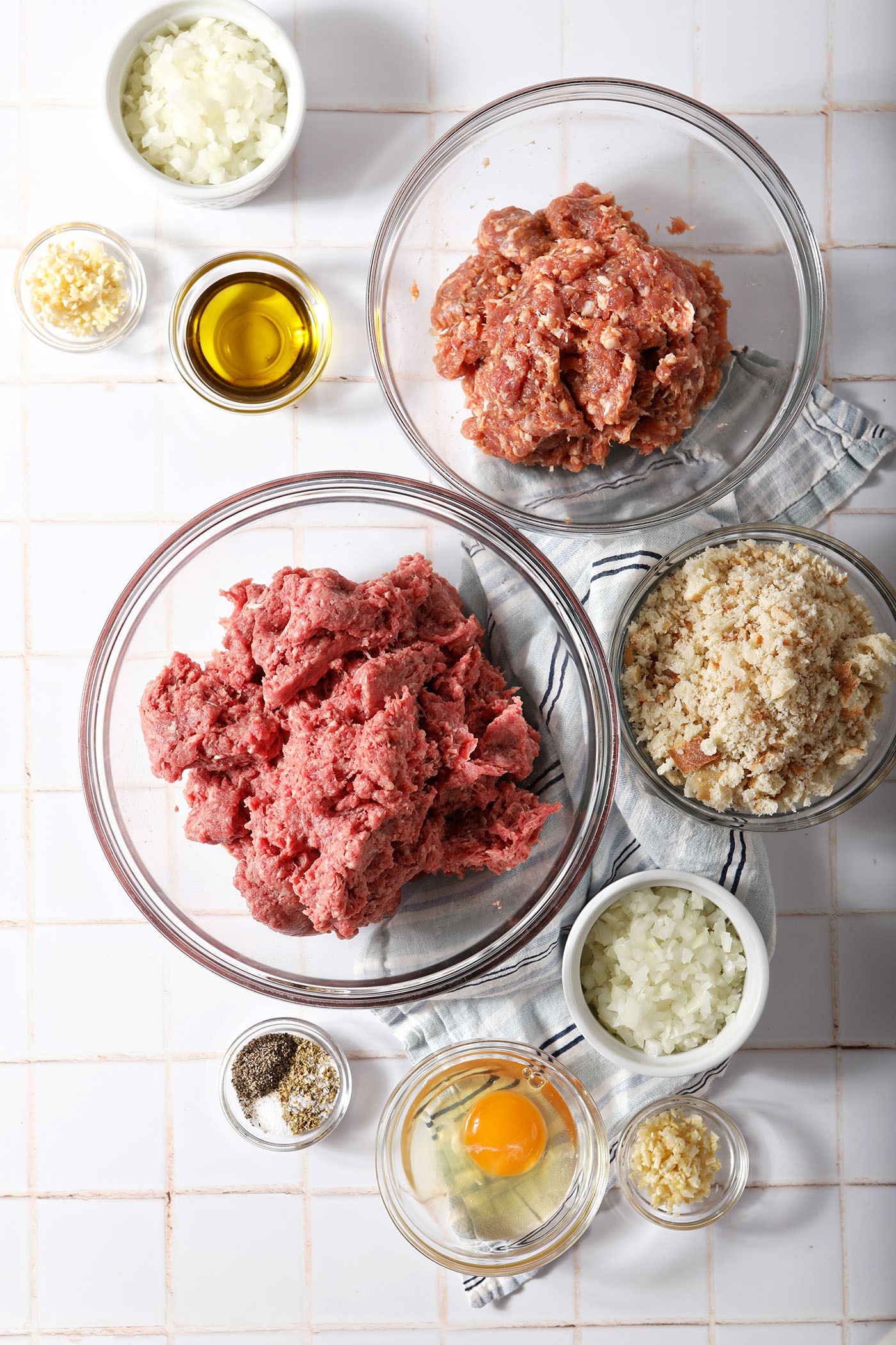 Overhead of ingredients in bowls to make beef and sausage meatballs on a white tiled surface