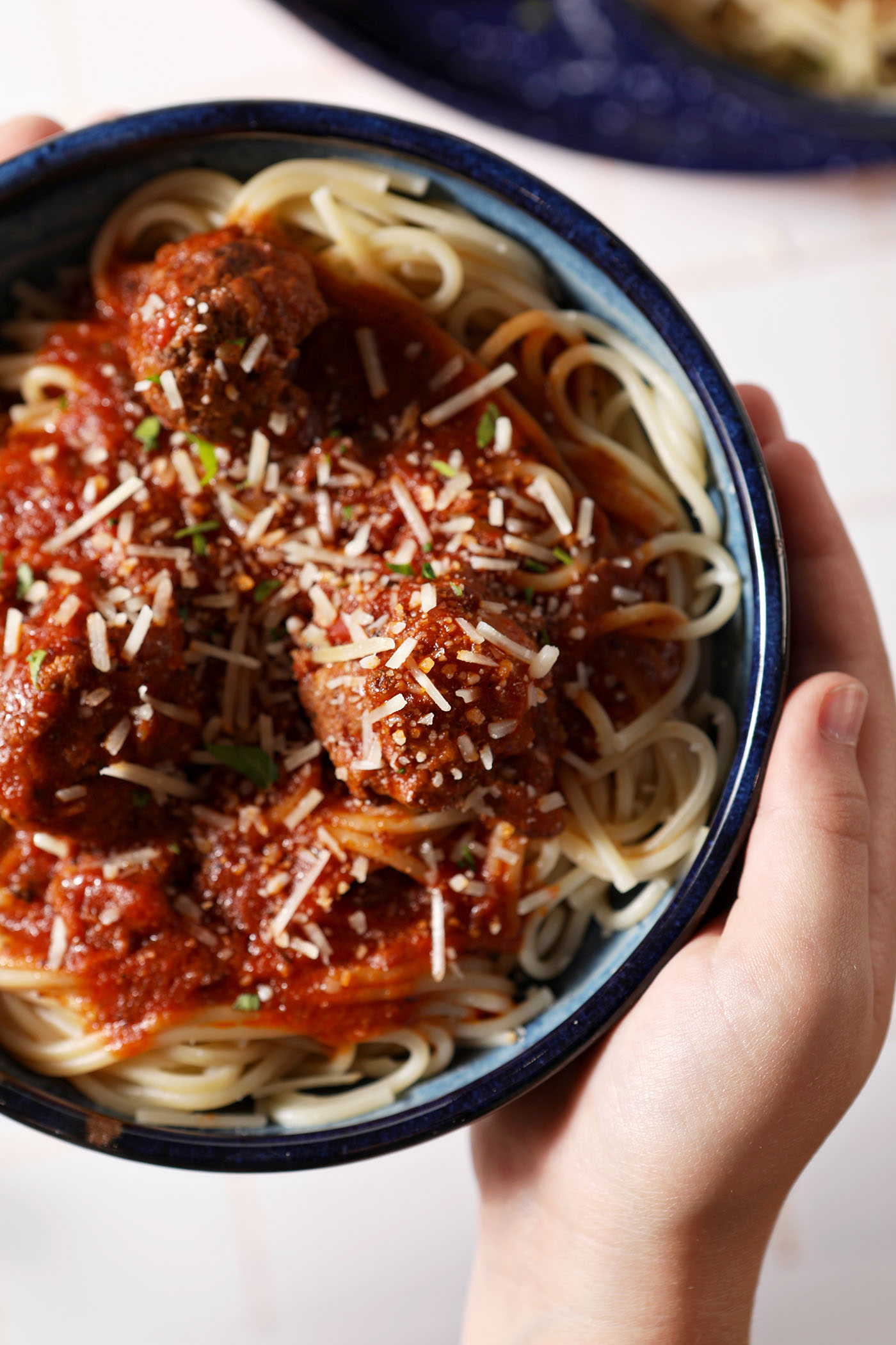 A person holds a blue bowl of spaghetti and meatballs, garnished with parsley and parmesan cheese