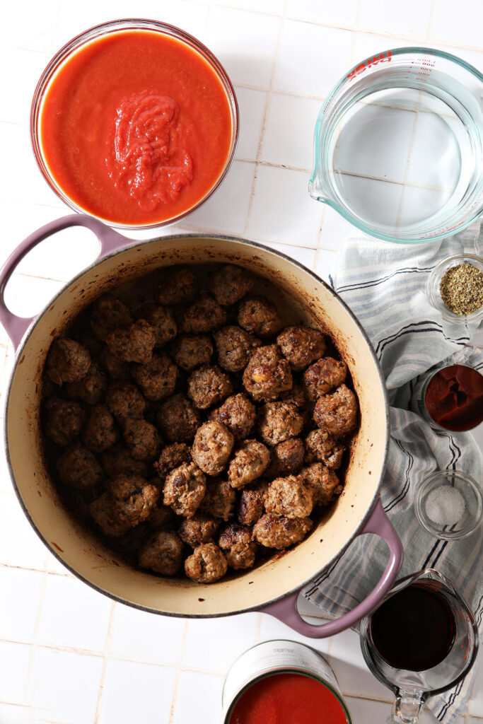 Cooked meatballs in a dutch oven surrounded by pasta sauce ingredients in cans and bowls