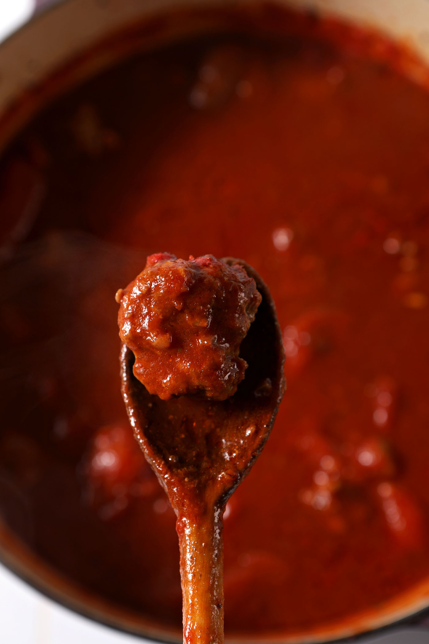 A wooden spoon holds a meatball above a pot of tomato sauce
