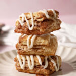 Three cinnamon biscuits topped with cream cheese frosting sit on top of each other on a plate on a pink surface