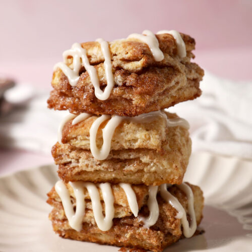 Three cinnamon biscuits topped with cream cheese frosting sit on top of each other on a plate on a pink surface