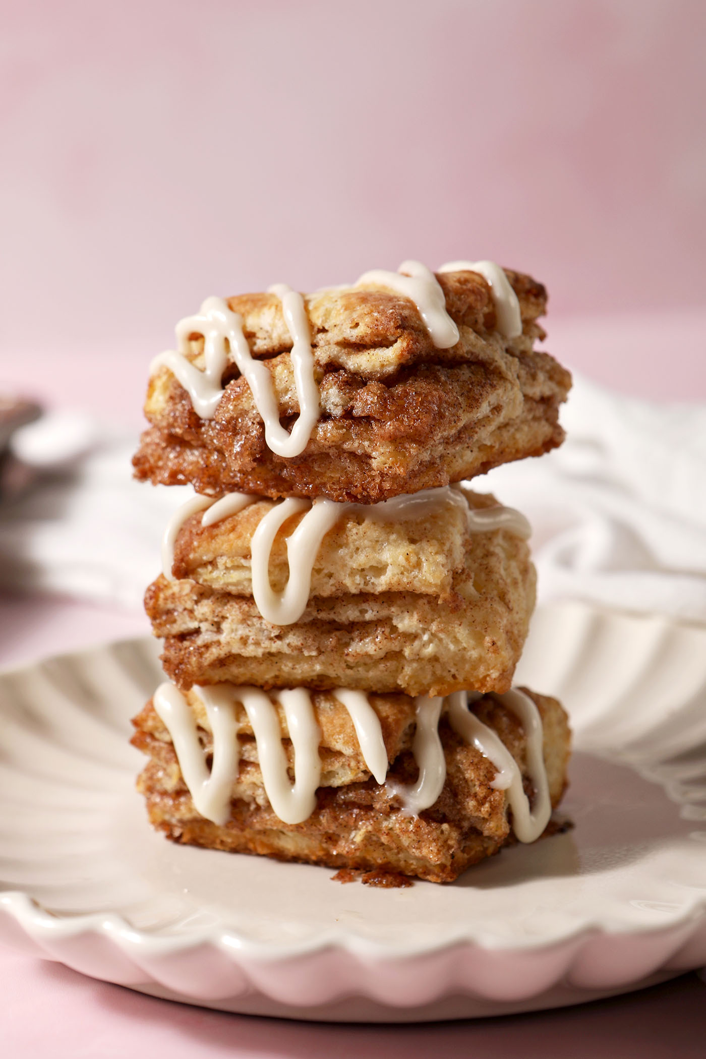 Three cinnamon biscuits topped with cream cheese frosting sit on top of each other on a plate on a pink surface
