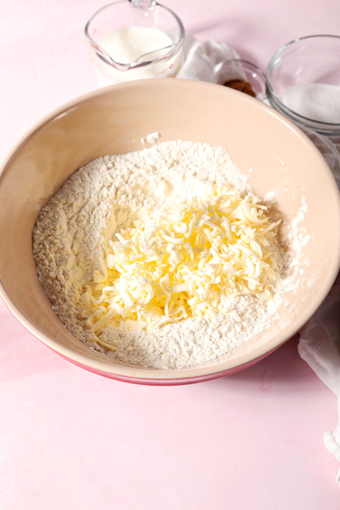 Shredded frozen butter sits on top of a flour mixture in a large bowl on a pink surface