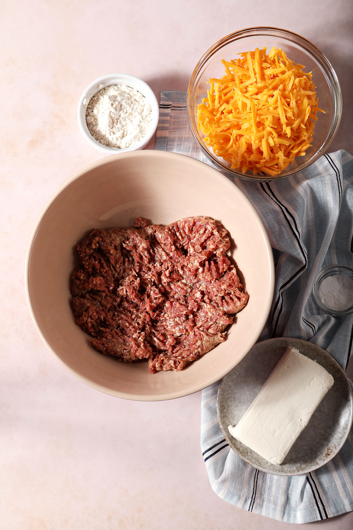Sausage and other ingredients to make sausage balls in bowls on a stone surface next to a blue and white striped linen