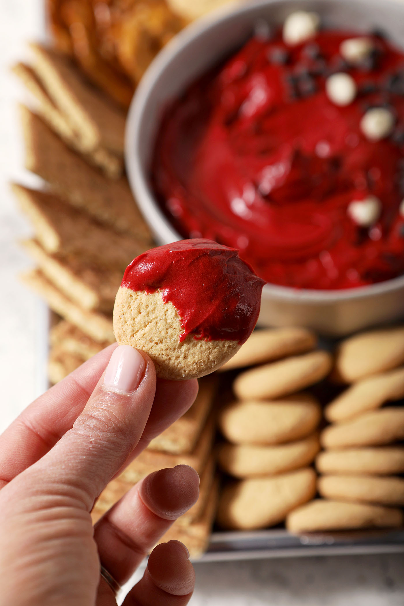A hand holds a dipped vanilla wafer cookie above red velvet dip above a platter