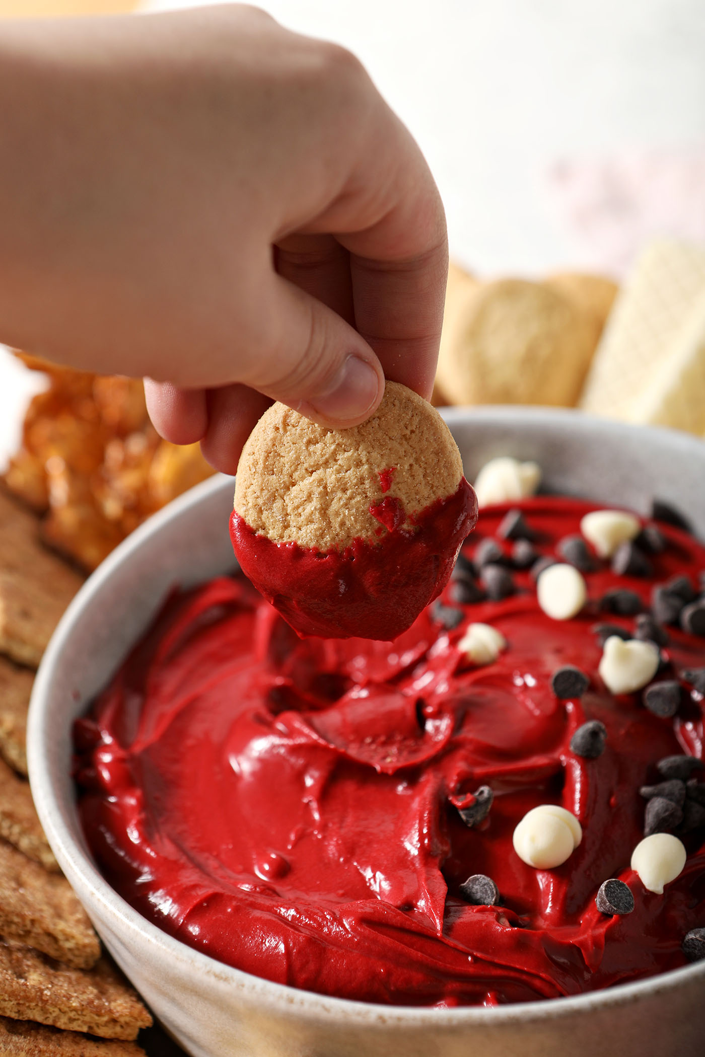 A hand dips a vanilla wafer cookie into red velvet cheesecake dip