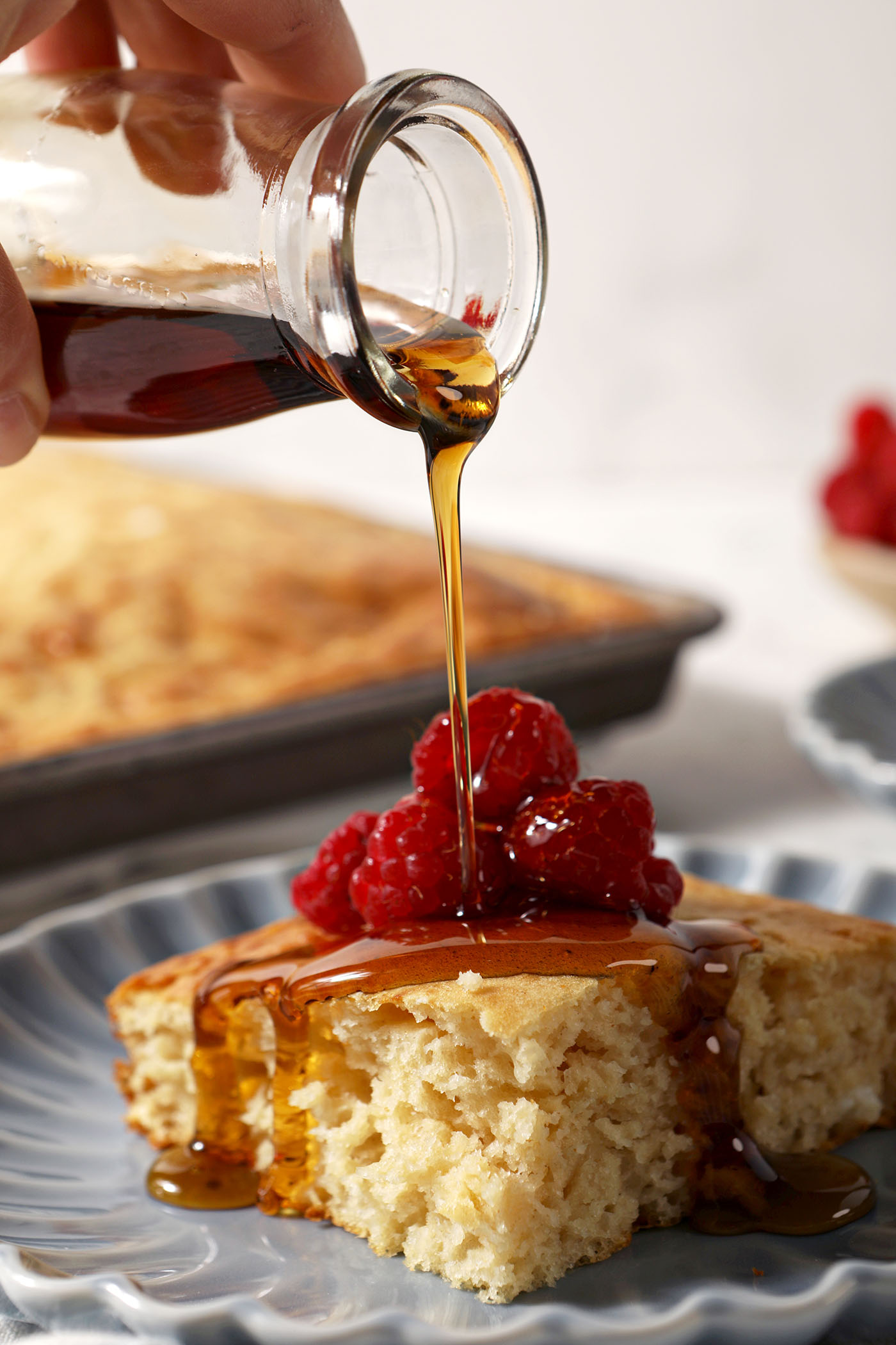 Syrup pours on top of a sheet pan pancake on a blue plate garnished with raspberries