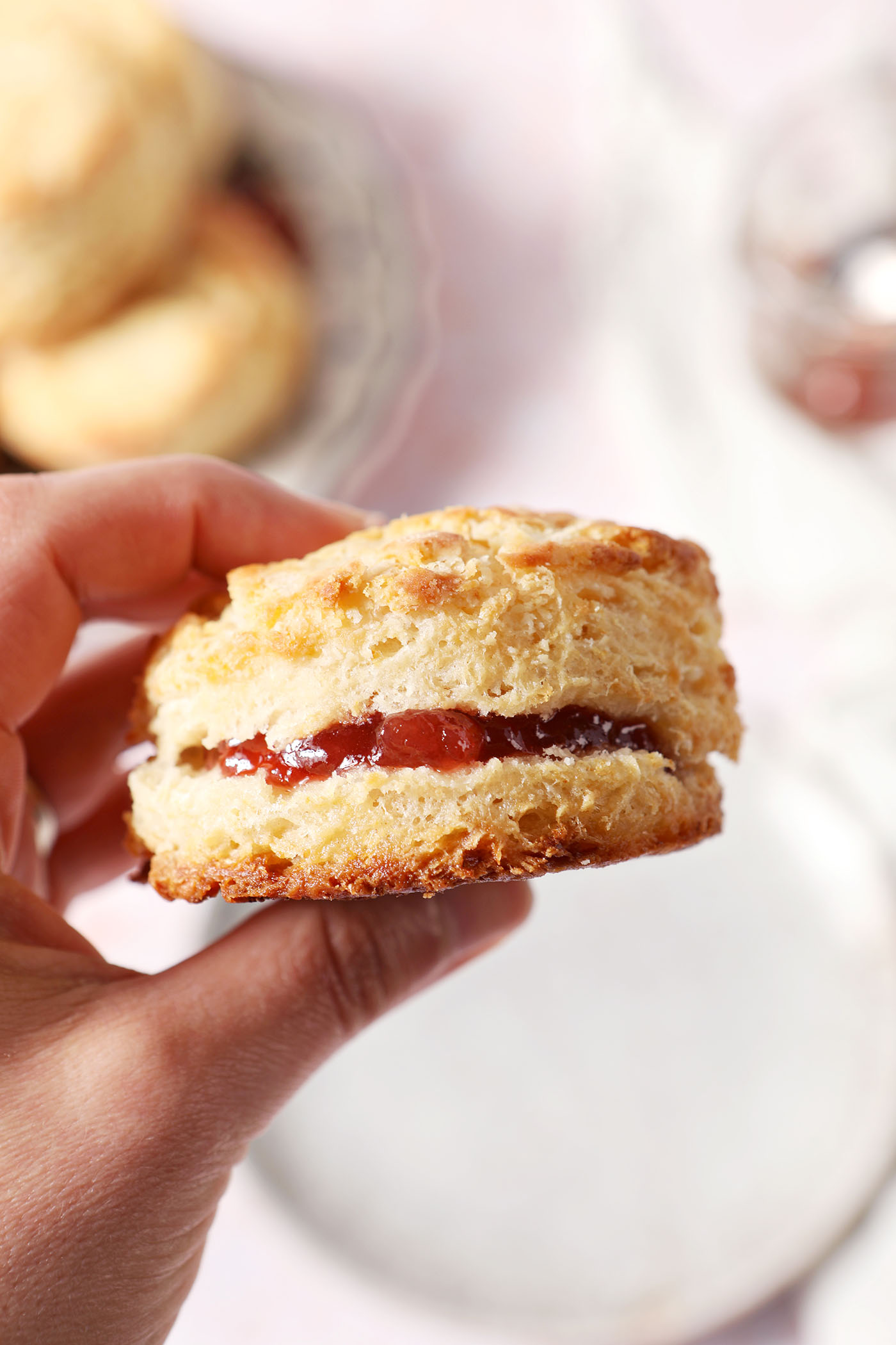 A hand holds a buttery biscuit filled with jam above a surface with more biscuits