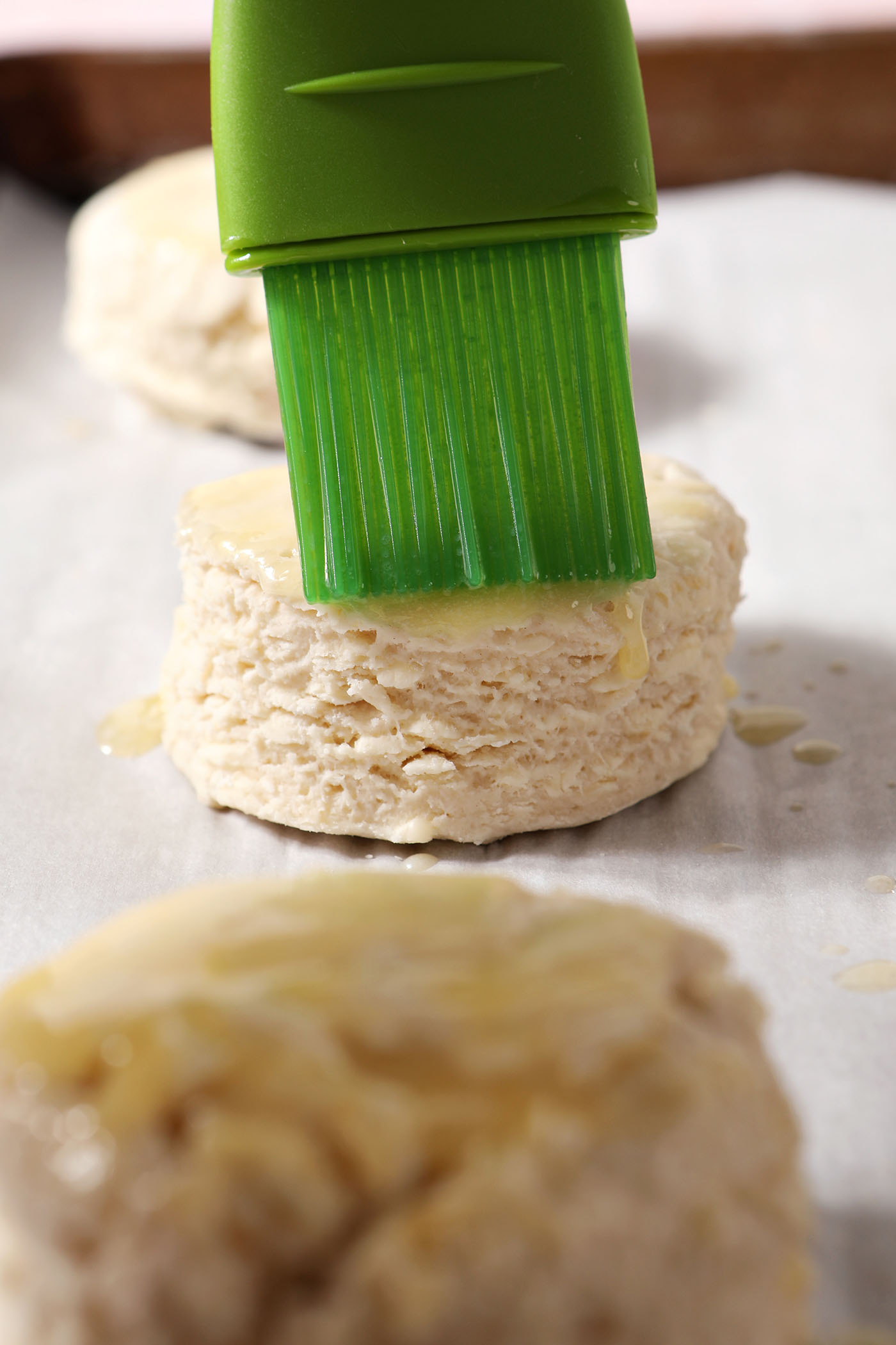 A green pastry brush brushes melted butter on top of frozen biscuits on a sheet pan