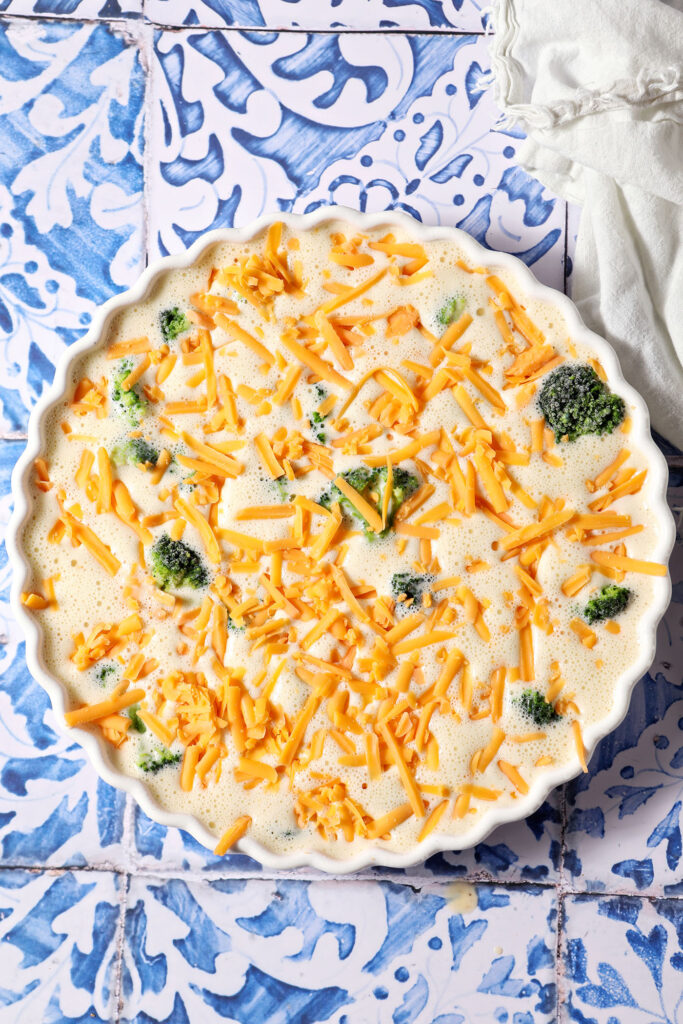 An unbaked quiche topped with bonus shredded cheddar cheese in a white tart pan on a blue and white tiled surface