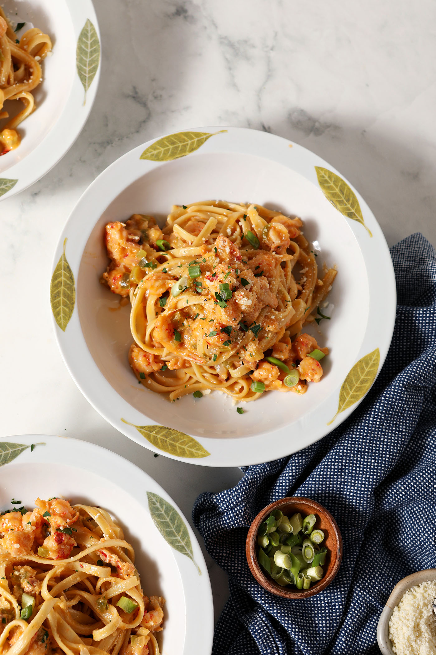 Overhead of three bowls of crawfish pasta garnished with green onions, parsley and parmesan on marble next to a blue linen