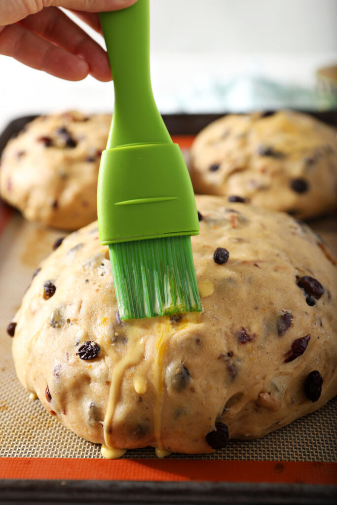A green pastry brush brushes an egg wash mixture over a boule of easter bread