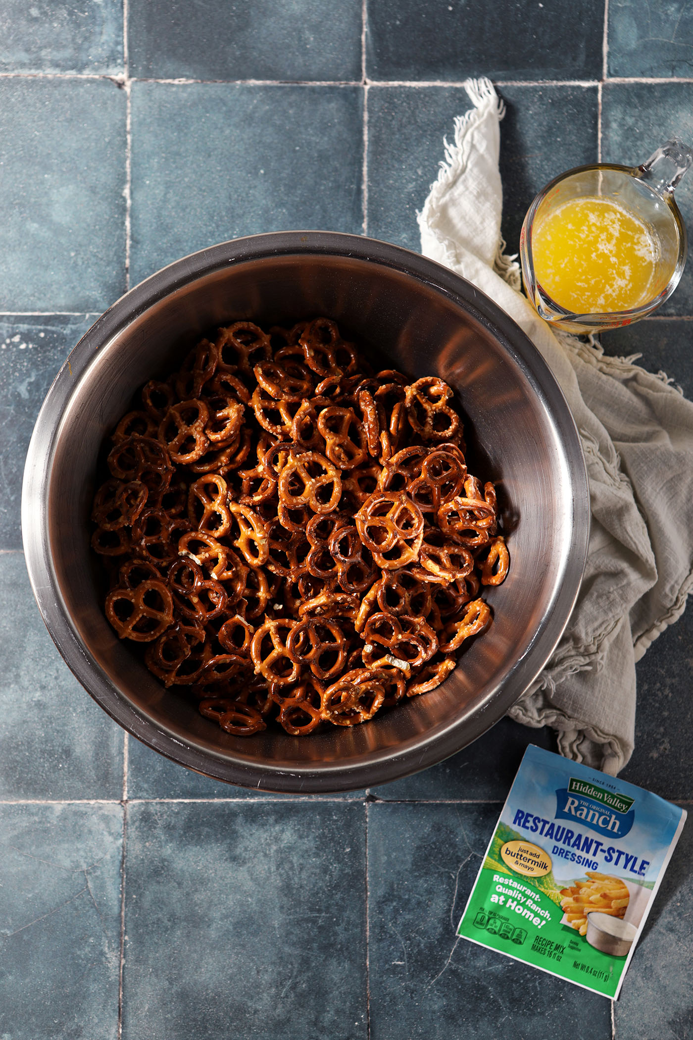 A bowl of pretzels with a packet of ranch dressing and a measuring cup of melted butter on a dark teal tiled surface