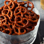 A glass bowl of ranch pretzels on a dark teal tiled surface in front of two cups of beer