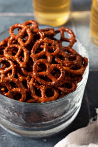 A glass bowl of ranch pretzels on a dark teal tiled surface in front of two cups of beer