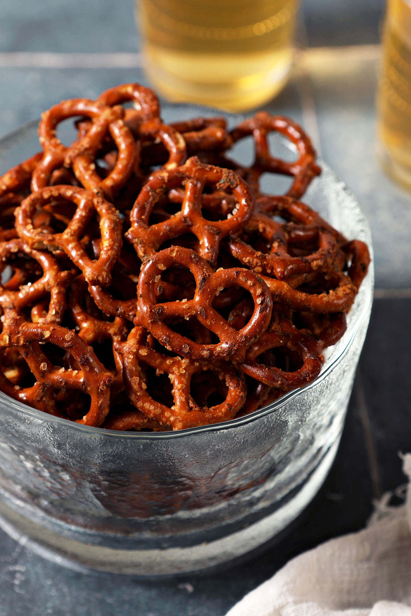 A glass bowl of ranch pretzels on a dark teal tiled surface in front of two cups of beer
