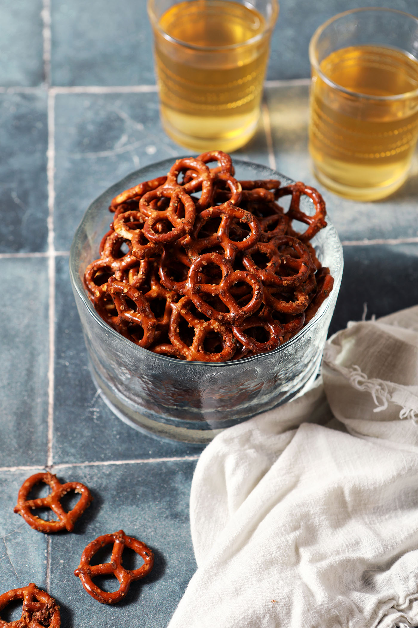 A glass bowl of ranch pretzels with some pretzels on a dark teal tiled surface with a white linen in front of two cups of beer