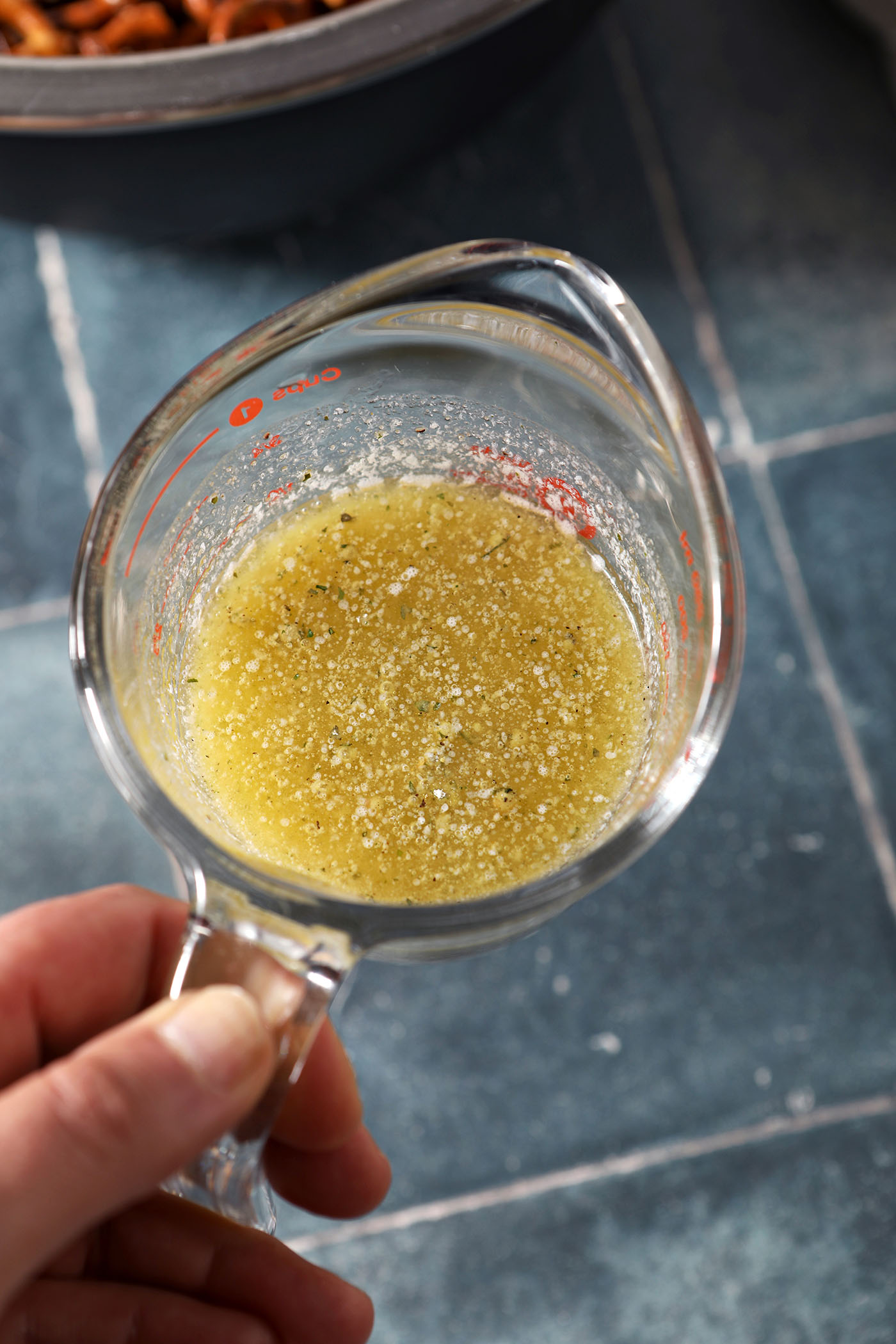 A hand holds a liquid measuring cup of melted butter and ranch dressing seasoning above a dark teal tiled surface