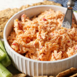 Side angle of a white bowl holding pimento cheese surrounded by crackers and celery sticks