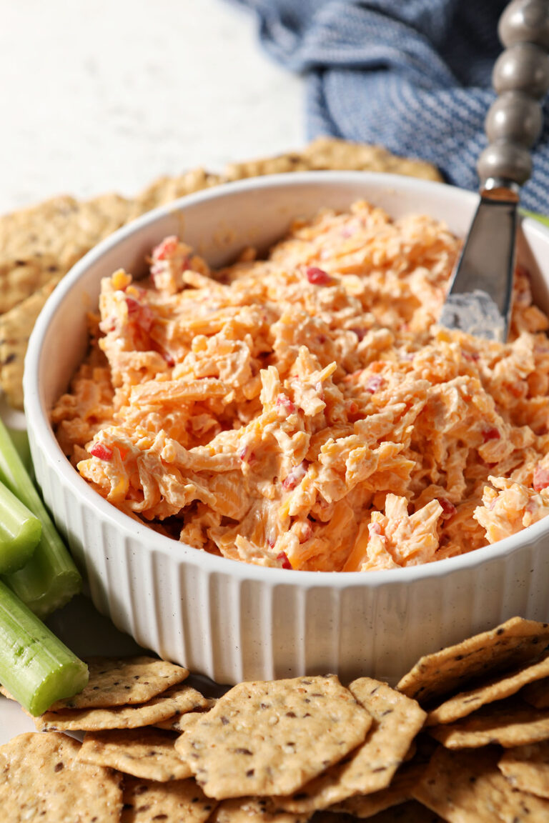 Side angle of a white bowl holding pimento cheese surrounded by crackers and celery sticks