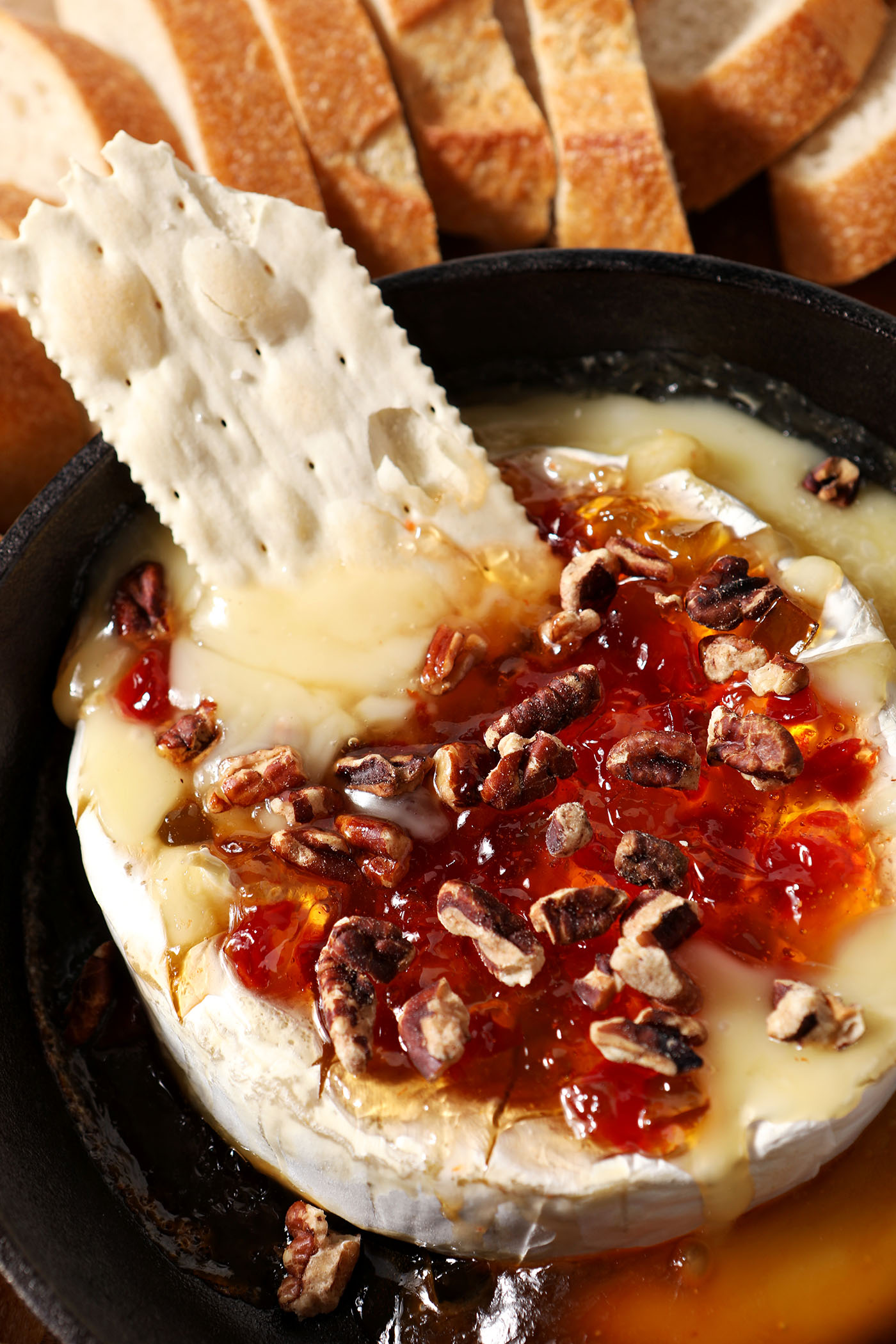 A cracker dips into gooey pepper jelly baked brie in a skillet next to baguette slices