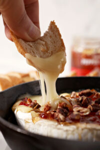 A baguette round is lifted out of a skillet holding Baked Brie with Pepper Jelly and Toasted Pecans
