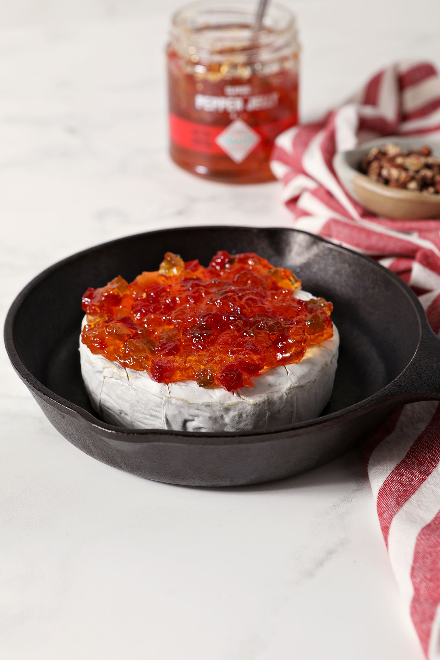 A wheel of brie in a small cast iron skillet topped with pepper jelly on a marble surface next to a red and white striped linen
