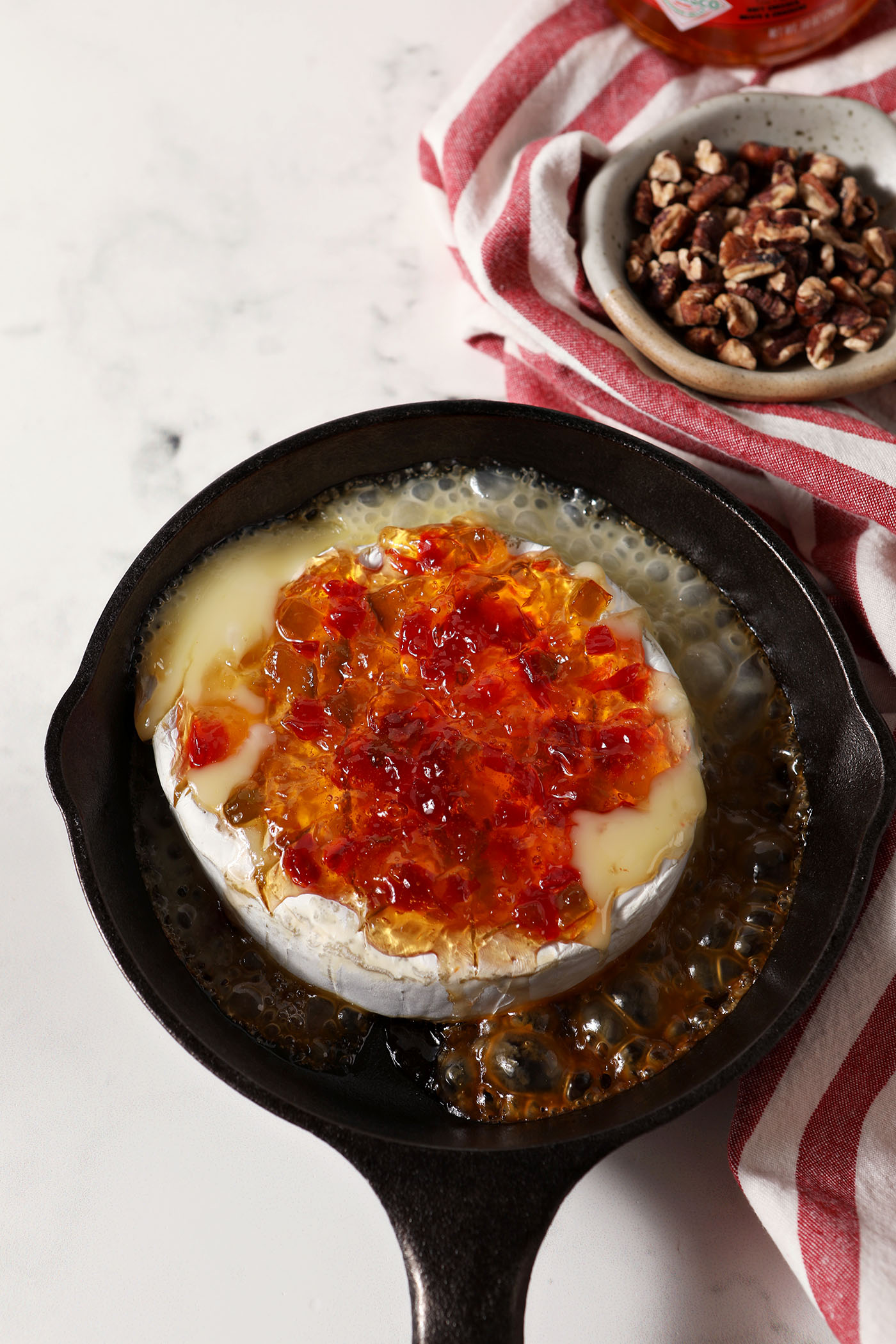 Overhead of a cast iron skillet holding a melty wheel of brie topped with pepper jelly on a marble surface next to a red and white striped linen