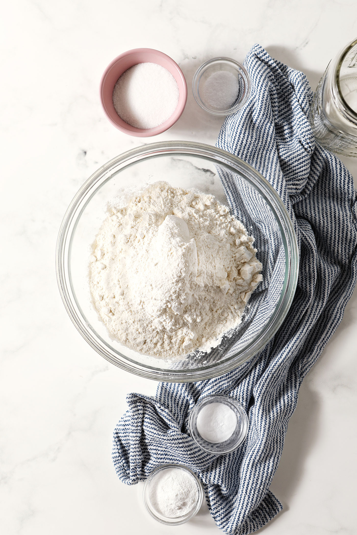 Overhead of bowls of dry ingredients to make homemade pancake mix recipe on a marble surface with a blue and white striped linen
