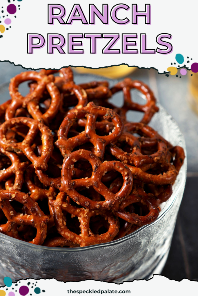 A glass bowl of ranch pretzels on a dark teal tiled surface in front of two cups of beer