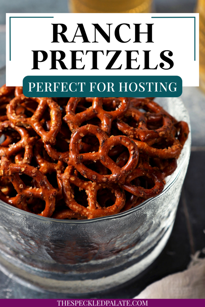 A glass bowl of ranch pretzels on a dark teal tiled surface in front of two cups of beer