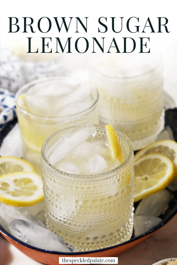 Four glasses of brown sugar lemonade on a blue tray with ice cubes and lemon wheels on a marble surface, next to a blue patterned linen, more lemon slices, and a bowl of brown sugar with the text "Brown Sugar Lemonade" on top.
