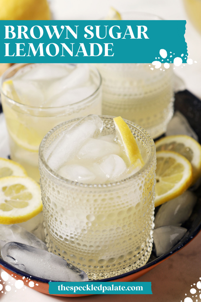 Four glasses of brown sugar lemonade on a blue tray with ice cubes and lemon wheels on a marble surface, next to a blue patterned linen, more lemon slices, and a bowl of brown sugar with the text "Brown Sugar Lemonade" on top.